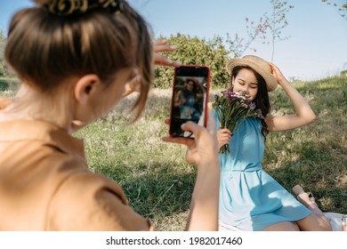 Back view of Young woman holding cell phone and making photo of her girl friend. Two girlfriends take pictures of each other on Smartphone camera on nature background. Summer holidays and technology - Powered by Shutterstock