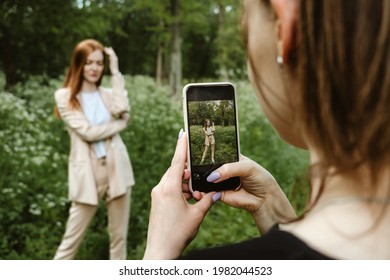 Back view of Young woman holding cell phone and making photo of her girl friend. Two girlfriends take pictures of each other on Smartphone camera on nature background. Summer holidays and technology - Powered by Shutterstock