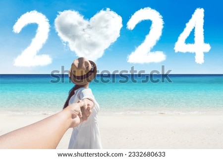 Back view of young woman holding hand of her husband while walking together toward 2024 new year numbers at beach