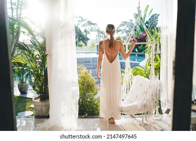Back view of young woman on terrace of resort hotel. Concept of tourism, vacation and weekend. Pretty girl with tattoos wearing dress. Sunny day. Idyllic and tranquil lifestyle on Bali island
