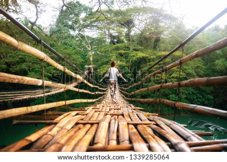 Back view of young woman on suspension wooden bamboo bridge across Loboc river in jungle. Vacation on tropical island. Bohol, Philippines