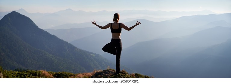 Back view of young woman performing yoga pose on grassy hill and looking at beautiful mountains. Sporty woman standing on one leg and doing Gyan mudra hand gesture while practicing yoga outdoors.