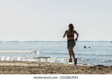 Back view of a young woman with a ponytail sprinting along the seashore on a sandy beach wearing shorts and a sports bra; a lady running by the sea on a sandy beach in sportswear on hot summer day
 - Powered by Shutterstock