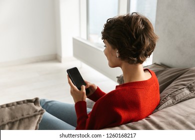 Back View Of A Young Woman Relaxing At Home, Listening To Music With Wireless Earphones, Holding Mobil Phone