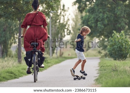Back view of young woman riding bike and boy near her riding skateboard performing cross feet tricks