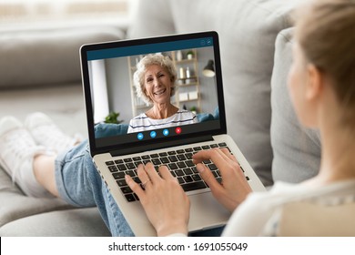 Back view of young woman sit relax on couch at home talk on video call with smiling mature mother, millennial girl rest have pleasant conversation with happy senior mom, use webcam on computer