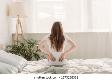 Back View Of Young Woman Sitting On Bed And Suffering From Back Pain