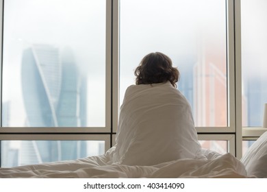 Back View Of Young Woman Sitting On Unmade Bed After Waking Up And Looking At City View In The Window. Female Model Relaxing Under Warm Blanket At Home. Copy Space