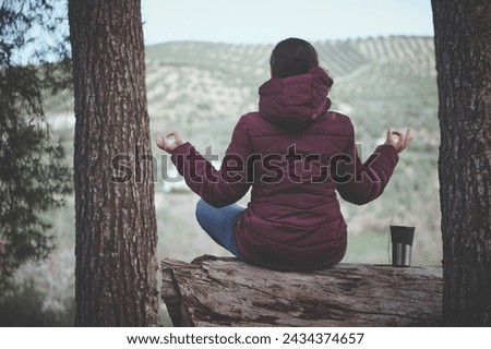 Back view of young woman sitting in lotus pose on a log in the forest, enjoying a meditation with the view of mountains on the background. Recreation. Lifestyle. Non urban scene. People and nature