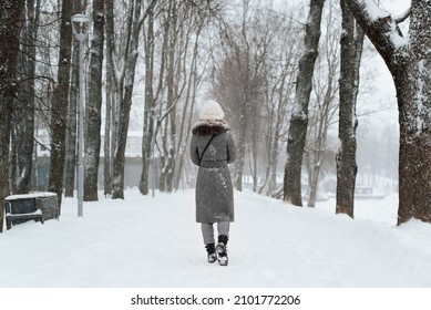 Back View Of Young Woman In Snow-covered Coat And Knitted White Hat Walking In Park In Winter, Outdoors. Rear View Of Lonely Girl Walking Along Snowy Alley.