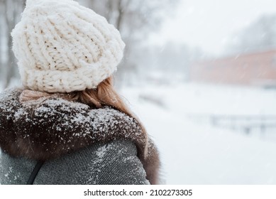 Back View Of Young Woman In Snow-covered Coat And Knitted Hat Enjoying Snowy Weather In Winter Park During Day, Copy Space. Rear View Of Lady On Nature Walk In Snowfall, Outdoors.