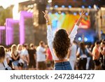 Back view of young woman at summer beach music festival. Raised hands with peace sign, enjoying live concert. Crowd, stage lights, sunset party vibes. Casual fashion, outdoor event, fun atmosphere.