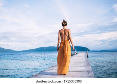 Back view of young woman with top knot in yellow dress walking on wooden pier against background of sea, mountains and clouds - Powered by Shutterstock