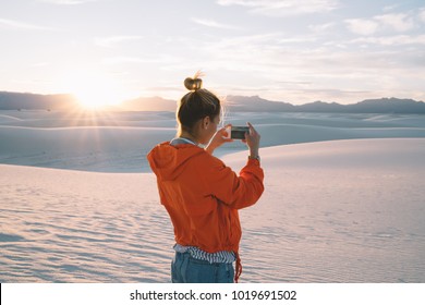 Back view of young woman using smartphone camera for making picture of sunset in white sand dunes at sunset, female traveler wanderlust taking photos on mobile phone during summer journey vacations - Powered by Shutterstock