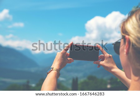 Back view of young woman using smartphone camera for making picture of Swiss Alps. Female traveler blogger taking photos on mobile phone during summer journey vacations.
