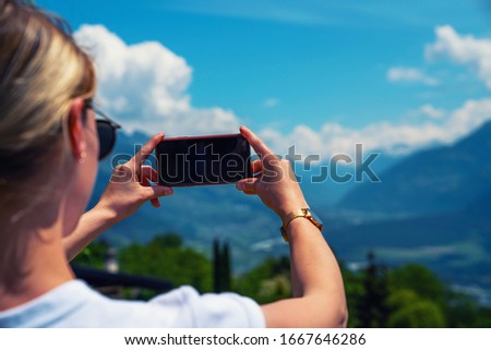 Back view of young woman using smartphone camera for making picture of Swiss Alps. Female traveler blogger taking photos on mobile phone during summer journey vacations.