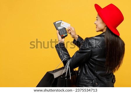 Back view young woman wear casual clothes red hat hold shopping paper package bags bank payment terminal process acquire credit card isolated on plain yellow background. Black Friday sale buy concept
