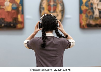 Back view of young woman wearing headphones and contemplates ancient arts. Student visiting gallery or museum. Concept of modern education and culture. - Powered by Shutterstock