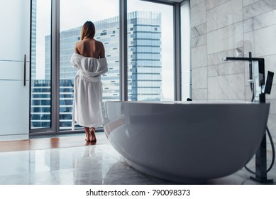 Back View Of Young Woman Wearing White Bathrobe Standing In Bathroom Looking Out The Window With Bathtub In Foreground