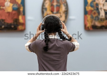 Back view of young woman wearing headphones and contemplates ancient arts. Student visiting gallery or museum. Concept of modern education and culture.