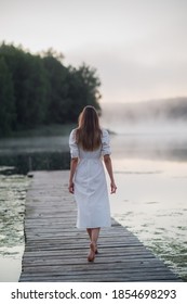 Back View Of Young Woman In White Dress Standing Alone On Footbridge And Staring At Lake. Foggy Chilly Morning With A Mist Over Water.