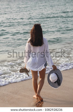 Back view of a young woman in a white beach outfit, holding a  white and blue straw hat walking on the beach. Travel & Summer vacation concept