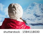 Back view of young woman in a winter knitted white cap, jacket and Christmas gloves drinking hot coffee or chocolate from red cup in the snowy mountains. 