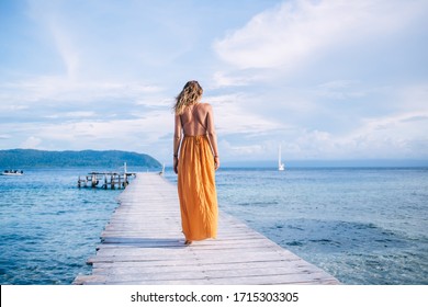 Back view of young woman in yellow dress walking on wooden pier against background of sea, mountains and clouds at sunny day - Powered by Shutterstock