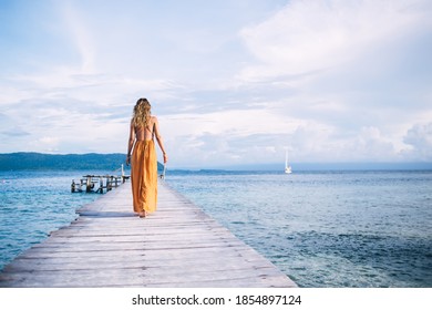 Back view of young woman in yellow dress walking on wooden pier against background of sea, mountains and clouds - Powered by Shutterstock