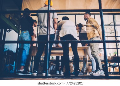 Back view of young working team working during meeting table sharing creative ideas and solutions, rear view through glass in coworking space of colleagues concentrated during brainstorming session