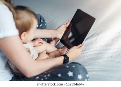 Back view.Close-up of tablet computer in hands of mother sitting with baby. Toddler girl sitting with mom on her lap, looking at screen of smartphone, watching cartoons, talking on video call.