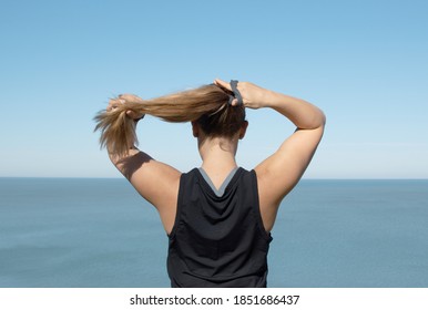 Back Of A Woman In A Black Athletic Shirt Sleeveless Putting Long Blonde Hair In A Pony Tail With Blue Water Blue Sky Background Looking Away 