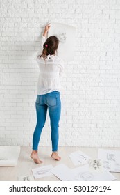 Back Of Woman Hanging Her Sketch On Wall. Female Artist Evaluate Her Painting Performance, Trying It On White Brick Wall, Free Space For Text