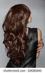 Back Of The Woman With Long Brown Curly Hair With Healthy Shine, Wearing A Leather Dress Over A Studio Background.