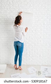 Back Of Woman Looking At Empty Paper On Wall Mockup. Artist Holding White Whatman Against White Brick Side Of Workshop, Free Space For Text Or Advertisement