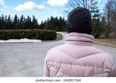 The Back Of A Woman Wearing A Pink Puff Parka And A Black Toque Standing On A Paved Driveway Lined With Coniferous Spruce Trees In The Countryside, March 2021, Ontario Canada. Blue Skies With Clouds. 