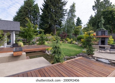 Back Yard Of A Contemporary Pacific Northwest Home Featuring A Deck A Spanning Creek-like Water Feature With A Landscaped Lawn And Custom Patio Fireplace In The Background.