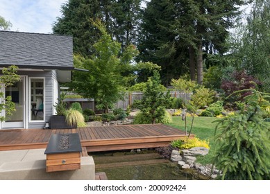 Back Yard Of A Contemporary Pacific Northwest Home Featuring A Deck Spanning A Creek-like Water Feature With A Landscaped Lawn In The Background.