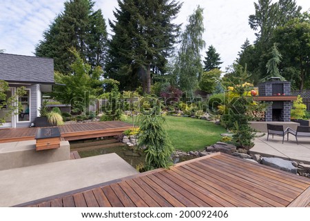 Back yard of a contemporary Pacific Northwest home featuring a deck a spanning creek-like water feature with a landscaped lawn and custom patio fireplace in the background.