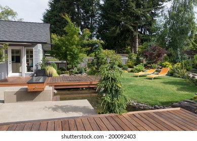 Back Yard Of A Modern Pacific Northwest Home Featuring A Deck Spanning A Creek-like Water Feature With A Landscaped Lawn And Two Inviting Lawn Chairs In The Background. 