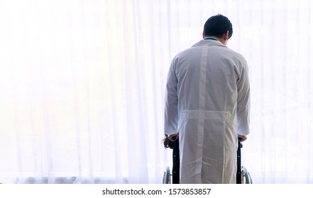 Back Of Young Doctor Wear White Coat Uniform Take Care Standing Push Patient Wheelchair While Looking Through The Window In Pain Room Ward At Hospital.