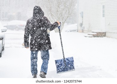 Back Of Young Man, Male Walking In Winter Coat, Cleaning, Shoveling Driveway From Snow On Street In Heavy Snowing Snowstorm, Holding Shovel, Residential Houses