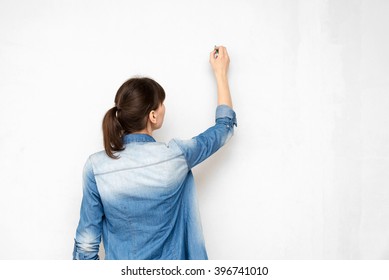 Back Of Young Woman In Casual Clothes With Pencil Drawing Or Writing On Empty Wall
