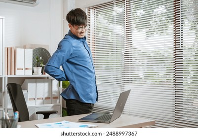 backache, executive, job, manager, missing, occupation, overworked, pain, papers, paperwork. A man in a blue shirt is hunched over a laptop computer. He is wearing glasses and he is in pain.