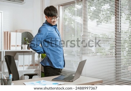 backache, executive, job, manager, missing, occupation, overworked, pain, papers, paperwork. A man in a blue shirt is hunched over a laptop computer. He is wearing glasses and he is in pain.