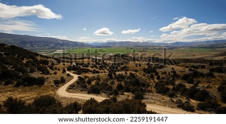 Backcountry nature panorama of road winding through dry rural landscape and green agriculture fields in background near Bendigo Central Otago South Island New Zealand