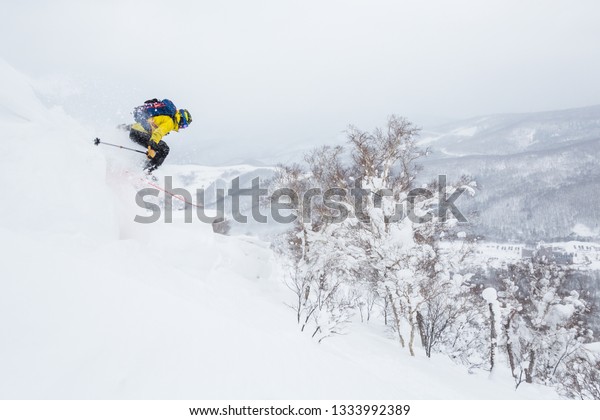 バックカントリースキーヤーは 日本の北海道の背景にある小さな山頂近くのコーニスからジャンプします 日本では 青いバックパックパウダースキー を行う黄色のジャケット コーニスエクストリームスキーを降りる の写真素材 今すぐ編集