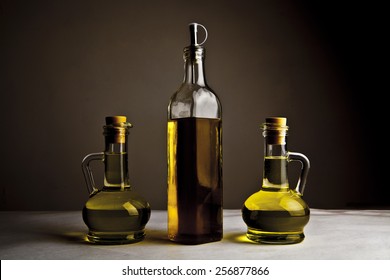 Backdrop Of Three Olive Oil Glass Transparent Bottle With Cork Stand On White Table On Kitchen Space Room, On Dark Black  Background Empty Space For Inscription 
