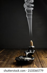Backflow incense on ceramic fountain on wooden table and white smoke on dark background