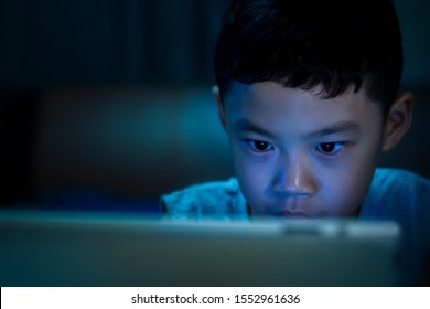 Background Of Asian Little Boy Age 6 Years Old Watching A Tablet And See Violence Content On Social Media With Dark Low Light In The Bedroom And Blue Lighting Reflection On His Face. Selective Focus. 